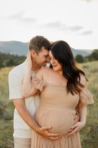 A pregnant wife hugging her husband in an intimate embrace. Surrounded by Colorado green grass and mountains. 