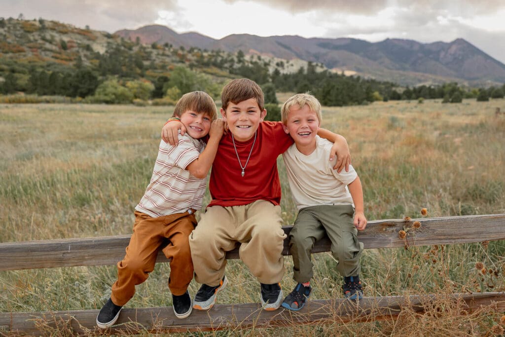 three children sitting on a bench, all smiling and snuggling for a posed portrait. Surrounded by Colorado Springs mountains and green grass. 