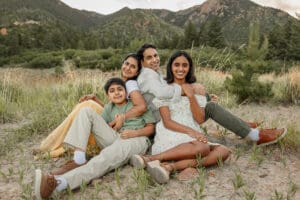 A family of parents and 2 children. They are sitting on the ground with their arms around each other smiling. Surrounded by green grass and mountains. Clients of pediatric chiropractor in Colorado Springs, Dr. Ryan.