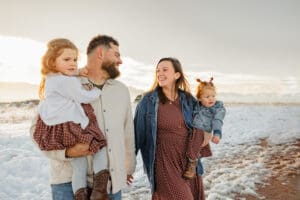 A family of parents and two small children. They are walking together, holding hands, and smiling at each other. Surrounded by Colorado Springs mountains and snow.